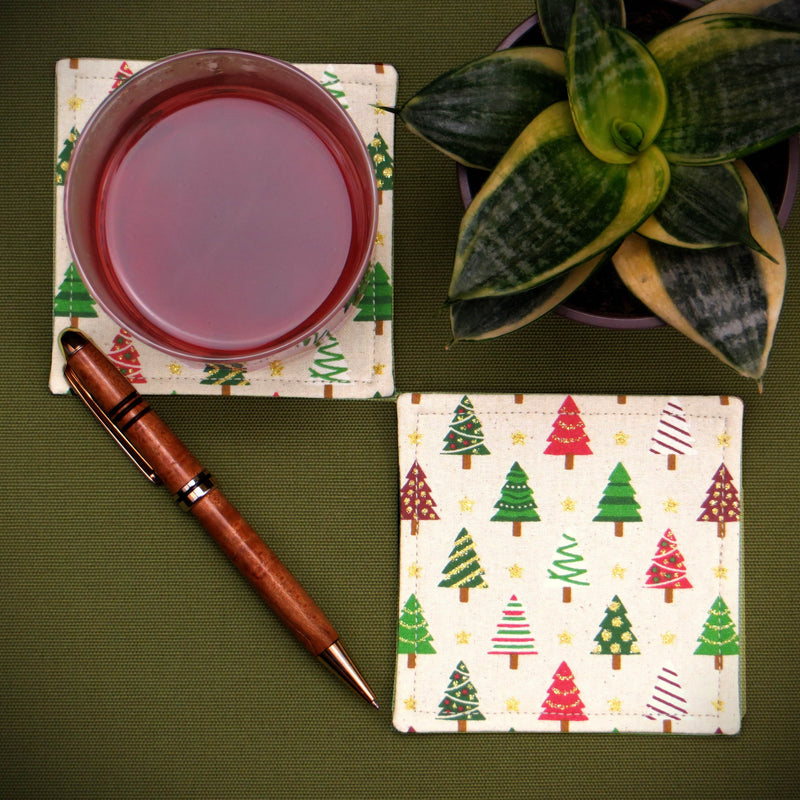 Set of two handmade cotton square coasters with a festive holiday design sitting on a green background with a pen, plant, and glass of cranberry juice
