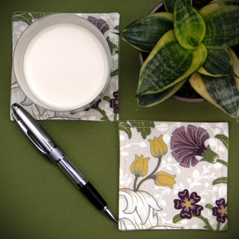 Pair of square fabric coasters with a white magnolia flower design with accents of green, yellow, and purple sitting on a table with a glass of milk