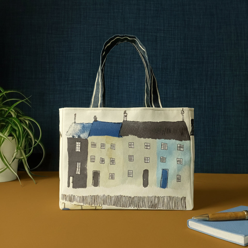 A handmade mini tote bag patterned with seaside houses and beach themed illustrations, and a pen with notebook and plant.