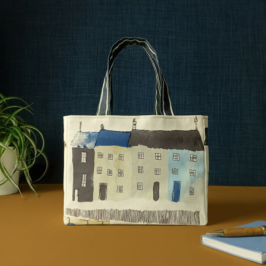 A handmade mini tote bag patterned with seaside houses and beach themed illustrations, and a pen with notebook and plant.