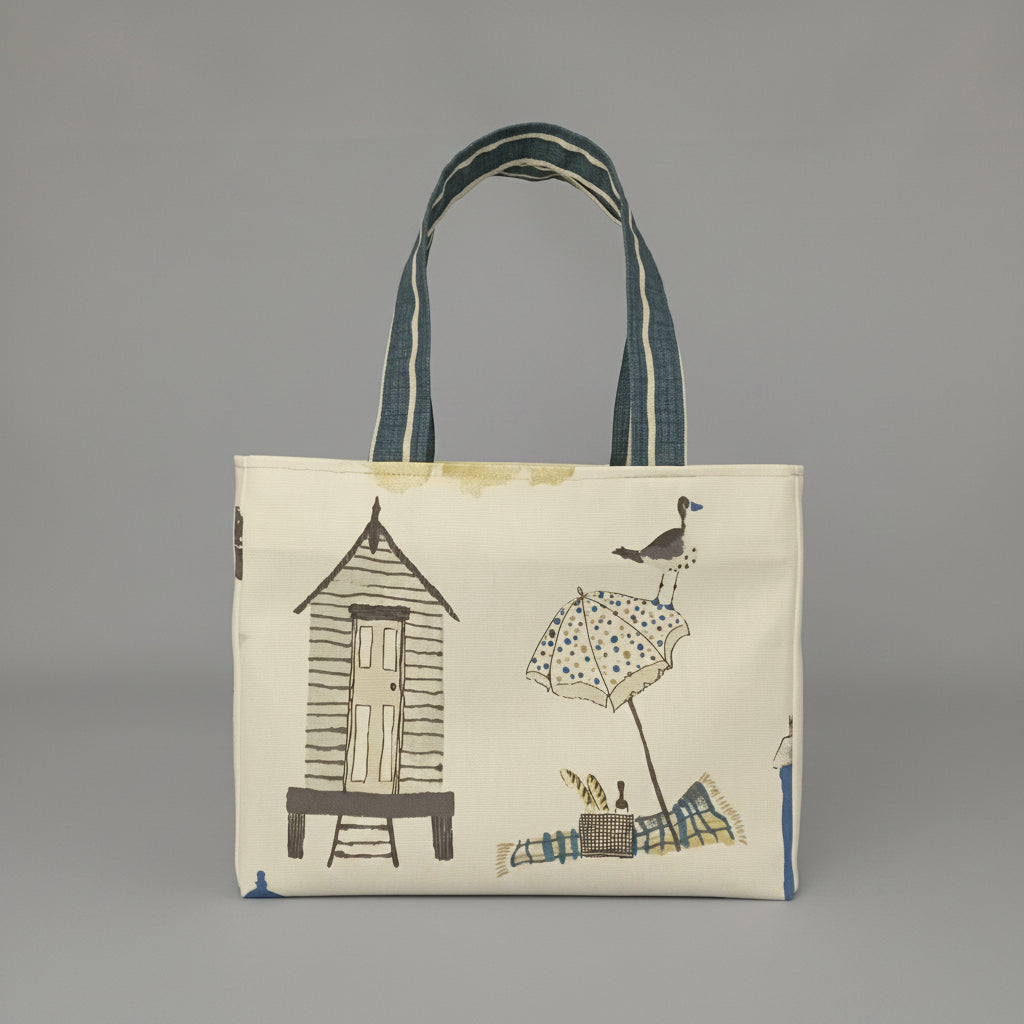 Back view of a handmade cotton mini tote bag patterned with a beach hut, beach blanket and beach umbrella illustrations on an off-white background.
