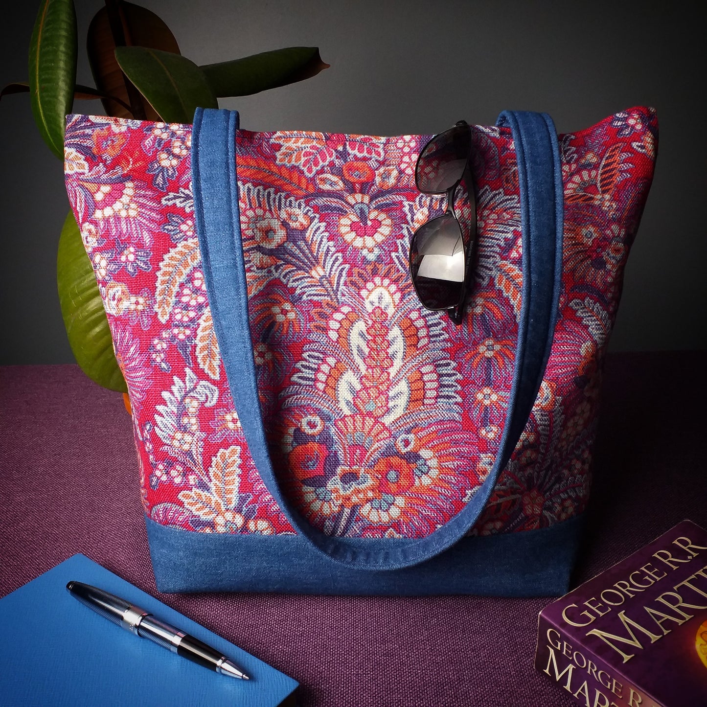 Large tote bag with multi-colour floral pattern, solid blue denim colour base sitting on a table with a notebook, pen, and plant