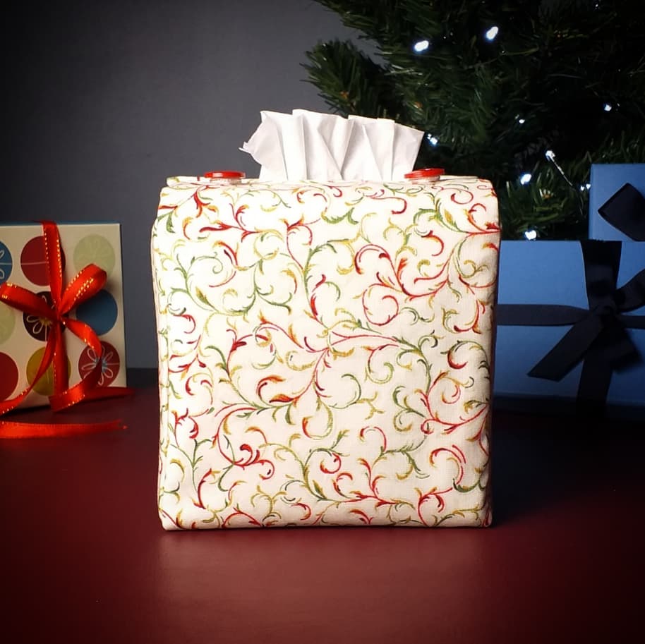 Square tissue box cover with holiday themed red and green swirls with metallic gold accent on cream colour background with Christmas themed decor