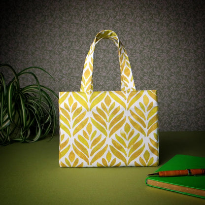Mini tote bag with yellow watercolour style leaf pattern on background sitting on a table with notepad, pen and plant